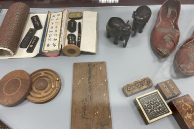 Różnorodne przedmioty kultury azjatyckiej są wyeksponowane w szklanej witrynie. Widać eksponaty takie jak figurki zwierząt, tradycyjne sandały, drewniane pudełka oraz naczynia stołowe. Przedmioty wykonane są z różnych materiałów, w tym drewna i ceramiki, i zdobione charakterystycznymi wzorami.