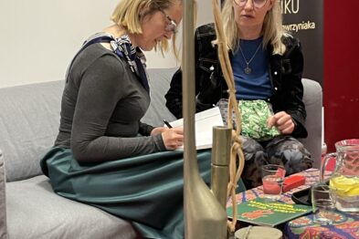 Dwie kobiety siedzą naprzeciwko siebie przy stole, jedna z nich podpisuje książkę. Na stole znajduje się stojak na lampę olejową, a w tle widać plakat z napisem 