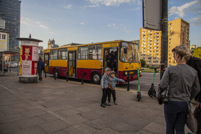 Ludzie spacerują po peronie kolejowym, gdzie wystawione są klasyczne żółto-czerwone autobusy. W tle widoczne są miejskie zabudowania i dworzec. Niebo jest jasne, co wskazuje na pogodny dzień.