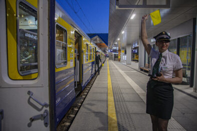 Mężczyzna w mundurze kolejowym macha żółtą chorągiewką na peronie stacji kolejowej obok żółto-niebieskiego pociągu. Jest wieczór, a na peronie widoczne są elementy oświetlenia. Osoba nosi ciemne okulary, czarny kapelusz i ma przy sobie przenośne radio.