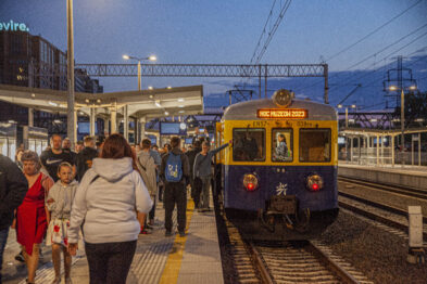 Stary pociąg pasażerski zatrzymał się na peronie; ludzie wysiadają z wagonów lub czekają na peronie. Jest zmrok, a oświetlenie peronu i pociągu tworzy uczucie wieczornej atmosfery. Na przedzie pociągu widać tablicę z napisem 