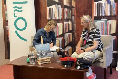 Dwie kobiety siedzą naprzeciwko siebie na krzesłach w bibliotece w trakcie spotkania czytelniczego. Za nimi widać półki z książkami, a między nimi umieszczony jest niski stolik z kilkoma egzemplarzami książki, notesem i butelką wody. Na pierwszym planie znajduje się roll-up z czytelnym tekstem w języku polskim.