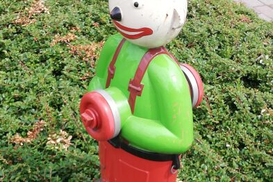 Przedstawia kolorową figurkę przedstawiającą ludzką postać, która jest przekształceniem urządzenia przeciwpożarowego. Figurka ma na sobie zielony strój z czerwonymi detalami, w tym kask, oraz żółte elementy na głowie i pasku. Ustawiona jest na zielonej powierzchni trawnika i zdaje się być ozdobą miejską o tematyce pożarniczej.