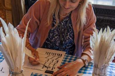Kobieta siedzi przy stole pokrytym w kratę obrusem i pisze piórem z atramentowego naczynia. Na stole leżą karty z wydrukowanymi ćwiczeniami kaligraficznymi, a obok stoją dwa szklane słoiki z piórami. Jest skupiona na pisaniu litery na papierze, uśmiechając się delikatnie.