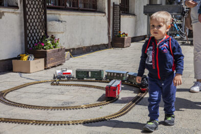 Dziecko stoi na zewnątrz budynku obok funkcjonującego modelu pociągu, który porusza się po okrągłym torze. W tle widać budynek i inne osoby. Model lokomotywy i wagon są kolorowe i szczegółowe.