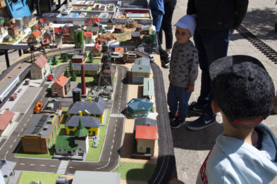 Dzieci wraz z dorosłymi oglądają makietę kolejową na zewnątrz podczas słonecznego dnia. Na makiecie widać modele budynków, drzew i pociągów. Osoby stojące po jednej stronie obserwują makietę z zainteresowaniem, a w tle widoczne są inne eksponaty kolejowe.