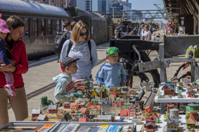 Dzieci wraz z opiekunami przeglądają różnorodne modele pociągów i kolejowych akcesoriów rozłożonych na stołach pod gołym niebem. Na stoisku widać miniaturowe budynki, drzewa i postacie, które tworzą makietę krajobrazu kolejowego. Tło sceny stanowią pociągi muzealne i otwarta przestrzeń stacji.