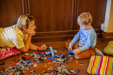Dwóch uczestników, kobieta i dziecko, siedzą na podłodze z kolorową poduszką i skupiają się na budowaniu konstrukcji z klocków LEGO. Na około nich rozrzucone są klocki LEGO w różnych kształtach i kolorach. Obydwoje wydają się być zaangażowani w tworzenie kolejowych modeli, odpowiednich do tematyki wydarzenia w muzeum.
