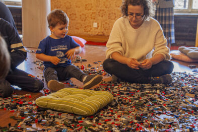 Dziecko w niebieskiej koszulce i kobieta siedzą na podłodze i bawią się kolorowymi klockami LEGO. Wokół nich na dywanie rozrzucone są mnóstwo małych elementów. W tle dostrzegalne są inne dzieci skupione na zabawie oraz jasne ściany pomieszczenia.