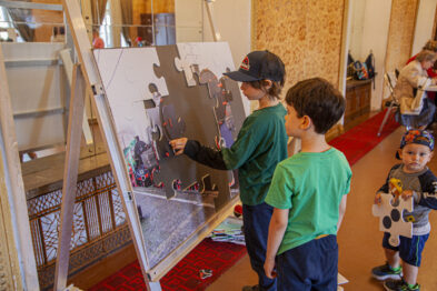 Dwóch chłopców w wieku przedszkolnym bawi się puzzlami z wizerunkiem lokomotywy umieszczonymi na stojaku. W tle widać innego chłopca i fragmenty wnętrza muzeum. Dzieci skupiają się na dopasowywaniu kształtnych elementów układanki.