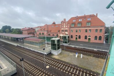 Czerwony neogotycki budynek dworca kolejowego z przełomu XIX i XX wieku dominuje na fotografii; jego dwuspadowe dachy i wysokie okna są charakterystyczne dla stylu architektonicznego. Przed budynkiem znajduje się peron oraz zestaw torów kolejowych, które biegną równolegle do dworca. Obok głównego budynku widoczne są także mniejsze konstrukcje, w tym klatka schodowa prowadząca na peron.