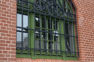Duże, łukowate okno o neogotyckich detalach zdobi ceglana ściana dworca kolejowego. Kunsztownie wykończona metalowa kratownica nadaje oknu elegancki charakter. Cegły są ułożone w regularnych rzędach, a okno jest zamknięte, przepuszczając światło do wnętrza.
