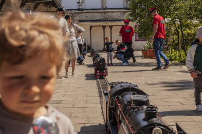 Dziecko obserwuje miniaturową lokomotywę parową, która porusza się po torach na zewnątrz. W tle widoczni są inni odwiedzający, w tym dorośli i dzieci, którzy także uwagę skupiają na modelu kolejowym. Słoneczna pogoda sprzyja atmosferze wydarzenia zorganizowanego na świeżym powietrzu w otoczeniu architektury muzeum.