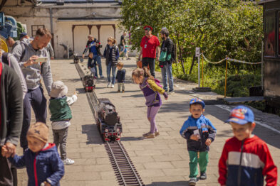 Na świeżym powietrzu dzieci obserwują i podążają za miniaturowym modelem pociągu, który przejeżdża po małych torach. Dorośli towarzyszą im, uważnie przyglądając się całej scenie. Całość odbywa się w przyjemnej, słonecznej atmosferze podczas wydarzenia tematycznego w muzeum.