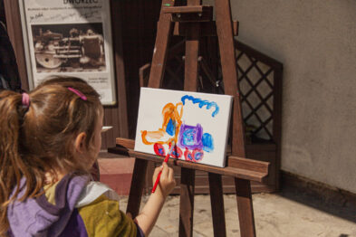 Dziecko maluje farbami na płótnie zamocowanym na drewnianej sztaludze. Obraz przedstawia kolorowe, abstrakcyjne formy. W tle widoczna jest częściowo ściana budynku i plakat z czarno-białą fotografią.