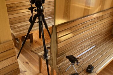 W drewnianej przestrzeni znajduje się statyw z kamerą ustawiony naprzeciwko drewnianej ławki, na której leżą dwa mikrofony z przewodami, a obok nich jest młotek. Ściany i podłoga wykonane są z jasnego drewna, a przez okna wpada delikatne światło. W pomieszczeniu widać też metalowe elementy konstrukcyjne, które dodają wnętrzu surowego charakteru.