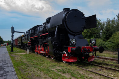 Pojazd parowy z czarnym lokomotywą i czerwonymi elementami stoi na torach kolejowych w dzień. Z przodu maszyny znajduje się okrągły reflektor, a kształty elementów są klasyczne dla dawnych pociągów. Oprócz lokomotywy widać także wagony pasażerskie oraz platformę peronową.