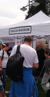 Grupa osób stoi przed białym namiotem z napisem "STACJA MUZEUM" i bacznie przygląda się czemuś lub słucha osoby, która nie jest widoczna na zdjęciu. Namiot znajduje się na otwartej przestrzeni z innymi obiektami w tle. Niebo jest zachmurzone sugerując możliwość niedługich opadów.