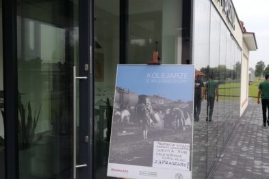 Na pierwszym planie stoi drewniany stojak z plakatem informacyjnym o Festiwalu Dziedzictwa Kresów. Plakat zawiera tekst i zdjęcie przedstawiające ludzi w strojach. W tle widoczne są otwarte drzwi szklanego budynku z napisem 