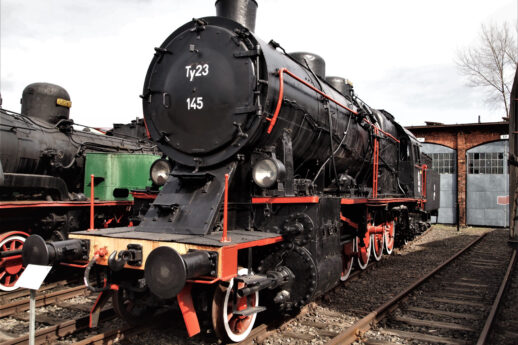 Czarno-czerwona, klasyczna lokomotywa parowa stoi na torach kolejowych; jest to maszyna z czasów, gdy parowozy były główną siłą napędzającą pociągi. Torowisko znajduje się obok innych lokomotyw i budynków kolejowych, które wydają się być częścią muzeum kolejnictwa. Niebo jest pochmurne, co dodaje zdjęciu lekko ponury, ale historyczny charakter.