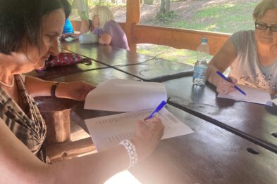 Grupa seniorów siedzi przy drewnianym stole w letnim otoczeniu, skupiając się na czytaniu i pisaniu na kartkach papieru. Stół otoczony jest zielenią, co sugeruje, że wydarzenie ma miejsce na zewnątrz, możliwe że w parku bądź lesie. Uczestnicy zaangażowani są w aktywność edukacyjną lub warsztatową, trzymając w rękach długopisy i materiały szkoleniowe.