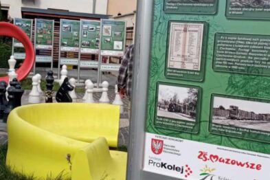 Stojak informacyjny z plakatami i zdjęciami przedstawia tematykę kolejnictwa, w tym mapy, stare zdjęcia i opisy. Obok tablicy znajduje się żółty stojak na ulotki. W tle widać miejski krajobraz z budynkami mieszkalnymi.