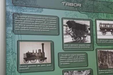 Panel informacyjny przedstawia historyczne zdjęcia i opisy dotyczące kolei, umieszczony na otwartej przestrzeni miejskiej. Widać fotografie parowozów, ludzi i budynków związanych z kolejnictwem. Tło panelu jest zielone, a treści zostały zorganizowane w tematyczne sekcje.