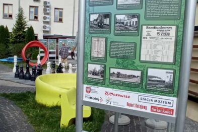 Stojak informacyjny z plakatami zawierającymi tekst i zdjęcia związane z kolejnictwem jest umieszczony na chodniku przed budynkiem. Obok stojaka widać składane stoły z żółtymi obrusami, na których leżą ulotki i inne materiały promocyjne. W tle znajduje się zielone drzewo i fragment budynku z białą elewacją.