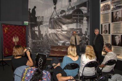 Grupa osób siedzi na krzesłach w pomieszczeniu z wystawą kolejową, obracając swoją uwagę w stronę mężczyzny stojącego obok historycznego sztandaru. Sztandar jest eksponowany w ozdobnej witrynie, a jego czerwone tło zdobi złocony orzeł i napisy. Duże, czarno-białe zdjęcia przedstawiające sceny kolejowe zdobią ściany sali, nadając wydarzeniu kontekst historyczny.