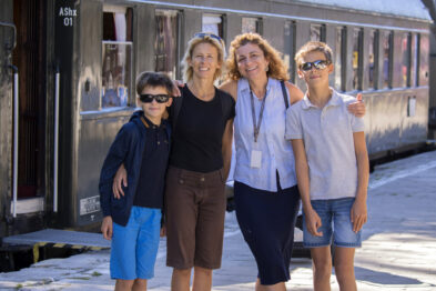 Na zdjęciu widoczni są cztery osoby – dwóch chłopców i dwie kobiety – stojących przed wagonami kolejowymi. Uśmiechają się oni do kamery, a słońce rozświetla scenę, rzucając cienie na peron. W tle widać historyczne wagony kolejowe, które są częścią ekspozycji muzeum.