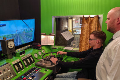 Dwóch mężczyzn skupia się na symulatorze lokomotywy, który reprodukuje kabinę maszynisty z wieloma przełącznikami i wskaźnikami. Na dużym monitorze wyświetlony jest widok z perspektywy maszynisty jadącej lokomotywy EU07. W tle widać eksponaty kolejowe i fragmenty wystroju wnętrza muzeum.