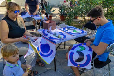 Zdjęcie przedstawia grupę osób, które biorą udział w zajęciach plastycznych na świeżym powietrzu. Dorosła kobieta i dziecko oglądają kolorowe prace położone na stole, podczas gdy mężczyzna pracuje nad malowaniem dużego, kolorowego numeru 