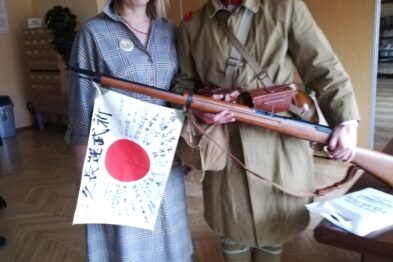 Na zdjęciu widać dwie osoby ubrane w historyczne kostiumy. Kobieta stoi po lewej stronie, ubrana w prostą sukienkę i trzyma flagę z czerwonym kołem na środku. Mężczyzna po prawej stronie jest ubrany w mundur wojskowy i trzyma w ręku drewniany karabin.
