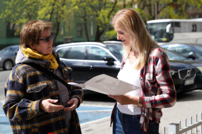 Dwie kobiety rozmawiają na zewnątrz przy słonecznej pogodzie; jedna trzyma otwarty folder lub kartkę. Obie stoją na chodniku, a w tle widoczne są zaparkowane samochody. Kobiety są ubrane w casualowe ubrania; jedna w szalik i kurtkę, a druga w kratowaną koszulę.