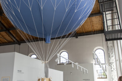Grupa osób stoi w przestronnym wnętrzu muzeum i patrzy na eksponat w kształcie balonu na ogrzane powietrze. Balon oznaczony jest napisem 