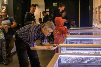 Grupa osób ogląda eksponaty umieszczone w szklanych witrynach w muzealnej przestrzeni. W centrum ujęcia mężczyzna skupia się na fotografowaniu jednego z eksponatów za pomocą telefonu komórkowego. Osoby w różnym wieku, w tym dzieci, przebywają w pomieszczeniu oświetlonym sztucznym światłem.