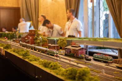 Makieta kolejowa jest umieszczona na długich stołach, przedstawiając krajobraz z torami, roślinnością i modelami pociągów. W tle widoczni są odwiedzający i uczestnicy wystawy, którzy obserwują i dyskutują o ekspozycji. W centrum uwagi jest model pociągu z zielonym wagonem, przemierzający makietę wzdłuż szczegółowo odwzorowanych torów kolejowych.