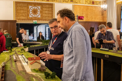 Dwie osoby obserwują detale makiety kolejowej prezentującej realistyczny teren z torowiskiem i zielonym krajobrazem. W tle inne osoby również zajmują się modelami kolejowymi. Makietę cechuje bogactwo detalów, w tym budowle i miniaturowe drzewa.