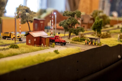 Makieta kolejowa przedstawia teren z budynkami, drzewami i miniaturą czerwonego samochodu. Prócz tego widnieją modele ludzi i sygnalizacja kolejowa. W tle rozciągają się zabudowania przemysłowe oraz fragment niebieskiego nieba.