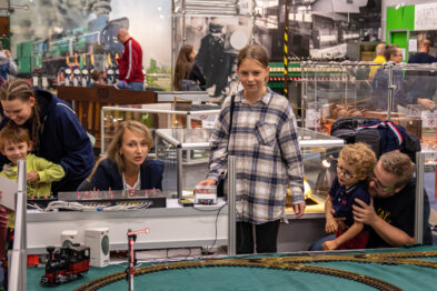 Grupa osób ogląda makietę kolejową prezentowaną na wystawie; dzieci stoja przy barierce, skupione na ruchomych modelach pociągów. W tle widać stoiska z modelarskim sprzętem i dodatkowymi akcesoriami. Centralnie umieszczony model kolejki przechodzi przez most - element ekspozycji otoczony jest przez zainteresowaną widownię.