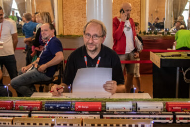 Osoby stoją i siedzą w pomieszczeniu z wysokim sufitem, obserwując makietę kolejową. Model pociągu z wieloma szczegółami jest umieszczony na torach makiety. Istnieje interakcja między uczestnikami, niektórzy noszą identyfikatory.