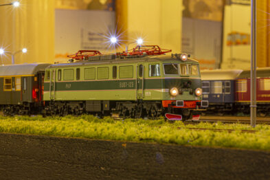 Model elektrycznego zielonego lokomotywy stoi na makiecie kolejowej z włączonymi światłami głównymi i pozycyjnymi. Obok lokomotywy widoczne są różnokolorowe wagony pasażerskie oraz towarowe. Makieta obejmuje także realistycznie wyglądającą trawę i żwir na torowisku.
