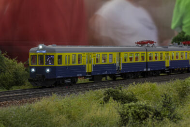 Model kolejowy przedstawiający skład pociągu, zbudowany z żółto-niebieskich wagonów, jedzie po szynach umieszczonych na zielonej makiecie. Otoczenie modelu tworzy niewielka ilość niskiej sztucznej roślinności, symulującej trawę i krzewy typowe dla krajobrazu kolejowego. W tle nieostro widać sylwetki ludzi, co świadczy o interaktywnej prezentacji modelu.