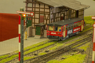 Czerwono-biały model pociągu stoi na torach obok makietowej stacji z imitacją muru pruskiego. Torowisko jest otoczone trawą i kilkoma niewielkimi postaciami ludzi. Poza semaforem w tle widoczne są inne tory oraz zielona przestrzeń.