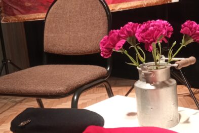 Na zdjęciu widać stół, na którym leżą dwa kapelusze kolejarza: jeden czarny i jeden czerwony z złotą wypustką. Za stołem stoi krzesło z oparciem, a na tle widać wiszącą tkaninę z polskim godłem – białym orłem w koronie na czerwonym tle. Na stole stoi również wazon z kwiatami, a całość kompozycji wydaje się być częścią wystawy lub wydarzenia kulturalnego.