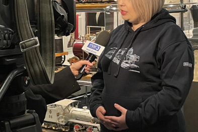 Kobieta udziela wywiadu przed kamerą telewizyjną wewnątrz muzeum kolejnictwa. Za nią widać gabloty z eksponatami związanymi z transportem kolejowym. Kobieta ma na sobie ciemną bluzę, a tło stanowią modele pociągów oraz kolejowe pamiątki.