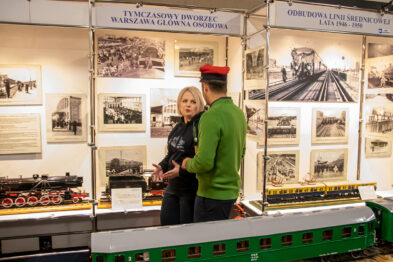 Na zdjęciu widać dwie osoby: mężczyznę w zielonej bluzie i czapce oraz kobietę w jasnym swetrze, którzy rozmawiają przy stoisku z eksponatami kolejowymi. W tle rozstawione są plansze z czarno-białymi fotografiami, przedstawiającymi motywy kolejowe. Pierwszy plan wypełniają modele pociągów ustawione na makiecie torów, w tym duży zielony model lokomotywy parowej.