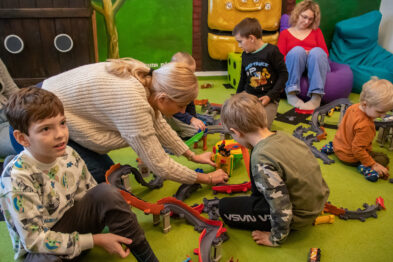 Dzieci bawią się kolorowymi torami kolejowymi i pociągami na podłodze. Osoba dorosła pomaga jednemu z dzieci w układaniu toru. W tle siedzi inna dorosła osoba na zielonym siedzeniu obserwująca zabawę.
