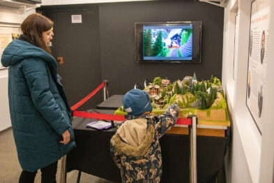 Kobieta i dziecko oglądają kolejową makietę oraz monitor wyświetlający film. Makietka zawiera modele zieleni oraz elementy kolejowe. Pomieszczenie, w którym się znajdują, to wnętrze muzealne z ekspozycjami i informacjami na ścianach.