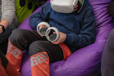Na fotografii widać dziecko siedzące na fioletowym worku sako, które ma na głowie zestaw do wirtualnej rzeczywistości (VR). Osoba ubrana jest w granatową bluzę i czerwone spodnie z odblaskowymi pasami; w rękach trzyma kontrolery VR. Wokół dziecka znajdują się inni ludzie, co sugeruje, że jest w miejscu publicznym, być może podczas wydarzenia związanego z kolei.